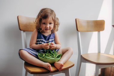 Küçük kız elinde yeşil bezelyelerle mutfakta oturuyor ve ipleri açmaya çalışıyor. Çocuk gri bir duvara yaslanmış bir sandalyede oturuyor ve taze bezelye yiyor.