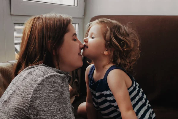 Pequena menina abraços e beijos sua mãe, filha abraça com a mãe em casa perto da janela Fotos De Bancos De Imagens