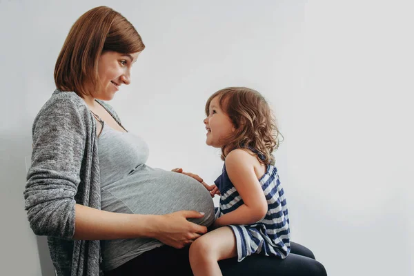 Barn flicka sitter på benen av en gravid mor och kommunicera med barnet i kvinnans mage Stockbild