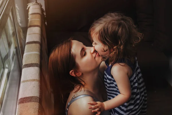 Liten flicka kramar och kysser sin mor, dotter kramar med mamma hemma nära fönstret Royaltyfria Stockfoton