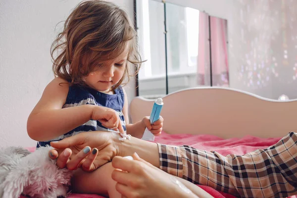 Bebê menina senta-se na cama no quarto e esfrega a perna creme, próximo senta-se mãe Imagens De Bancos De Imagens Sem Royalties