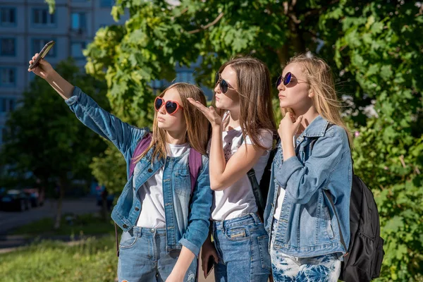 Tři dospívající dívky v létě venku. Díky fotografii osoby na telefonu. Nosí oblečení stylové džíny. Sluneční ochranné brýle. Dospívající dívky jsou blondýnky s batohy za jejich zády. — Stock fotografie