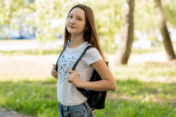 Beautiful teen girl in summer in a park outdoors in shade of a tree. Dressed in casual clothes. Behind the backpack. The schoolboy is going to school.