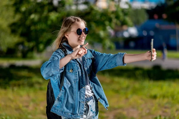 Adolescente en été dans un parc à l'extérieur. Dans les lunettes de soleil dans les mains tenant le téléphone fait une photo de personnes. Communique par appel vidéo. Une journée ensoleillée . — Photo