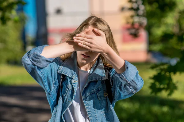 A little teenage girl covered her eyes with her hands. In the summer in the open air. A bright sunny day. The concept of reluctance to see. Very bright sun. The game of hide and seek After school. — Stock Photo, Image