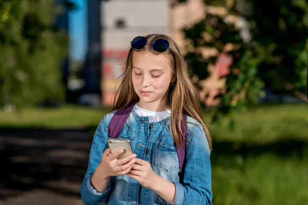 Una adolescente está cogida de la mano con un teléfono. Feliz sonrisa es escribir en las redes sociales. En verano al aire libre. Brillante día soleado en la cabeza de las gafas de sol . —  Fotos de Stock