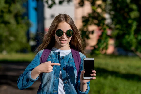 Una adolescente en un día soleado brillante con gafas de sol. En el verano en el parque al aire libre. En sus manos tiene un smartphone. Con un gesto de su mano señala el teléfono con el dedo . —  Fotos de Stock