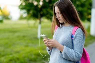 Küçük kız liseli esmer. Okulda okuldan sonra. Arkasından pembe bir sırt çantası var. Elinde bir akıllı telefon tutar. Film izlerken müzik kulaklık dinler. Ebeveynler arkadaşlarıma mesaj yazıyor.