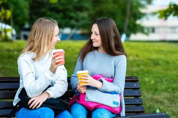 Gadis sekolahan di bangku. Teman-teman terbaik di taman musim panas di udara segar. Dia memegang kopi panas dan teh. Siswa wanita sepulang sekolah berlibur . — Stok Foto