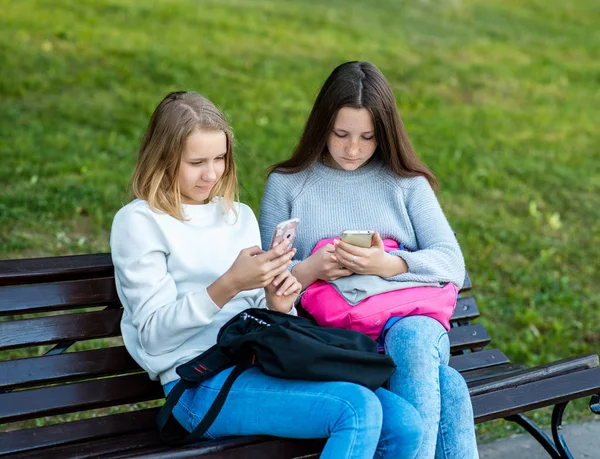 Nejlepší přátelé v létě v parku čerstvého vzduchu. Holky školačka na lavičce drží chytré telefony. Odpovídala v sociálních sítích. — Stock fotografie