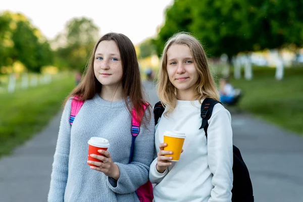 Две красивые девочки школьницы. Летом за городом на свежем воздухе. Он держит в руках горячий чай и кофе. Счастливая улыбающаяся поза перед камерой. Он отдыхает после школы. В летнем тени деревьев . — стоковое фото