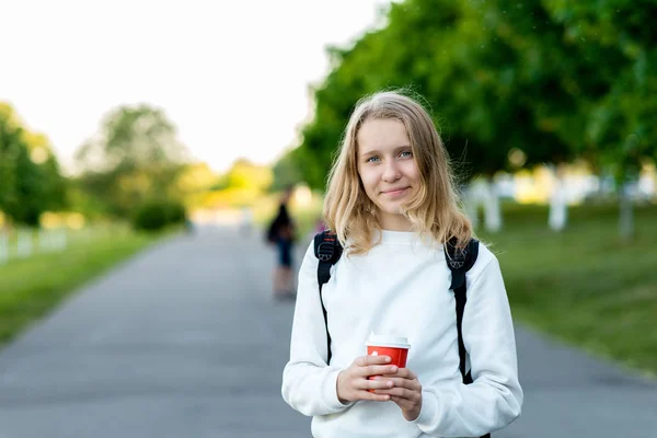 漂亮的女学生金发女郎。夏天在户外的女生。他拿着一杯热咖啡或他的手喝茶。微笑着在相机上摆姿势. — 图库照片