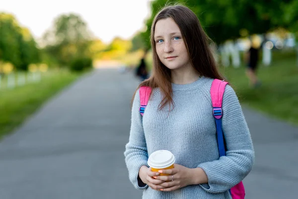 Dziewczynka jest nastolatkiem. Lato w przyrodzie. W ręku trzyma szklankę gorącej herbaty lub kawy. Wolna przestrzeń dla tekstu. Z plecaka na ramieniu po szkole. — Zdjęcie stockowe