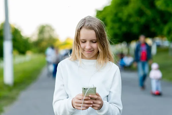 少女女子高生金髪。自然の中の夏。手に携帯電話で音楽を聞くスマート フォンを保持します。ソーシャル ネットワークで通信します。幸せな笑みを浮かべてください。市のにぎやかな通り。両親に Sms を書く. — ストック写真