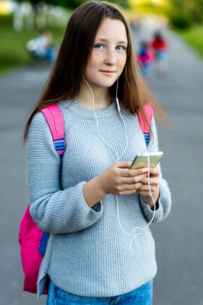 美しい少女ブルネットの女子高生。自然の中の夏。背中の後ろに、ピンクのバックパックです。ヘッドフォンと携帯電話の手。美しい青い目。そばかすの顔。髪は長い。幸せな音楽を聴く. — ストック写真