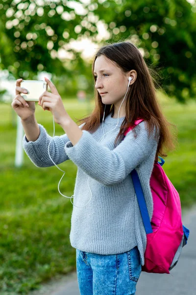 Ένα εφηβικό κορίτσι. Το καλοκαίρι στη φύση. Μελαχρινή με φακίδες στο πρόσωπό της. Κάνει μια φωτογραφία στον δρόμο. Πίσω από την πλάτη είναι ένα ροζ σακίδιο. Παίζουν μουσική στα ακουστικά. — Φωτογραφία Αρχείου