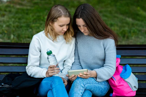 Две девушки-студентки. Лето на природе. Они сидят на скамейке и смотрят фильм в смартфоне. Держит в руках бутылку воды. Смотрел видео по телефону. Отдых после школы . — стоковое фото