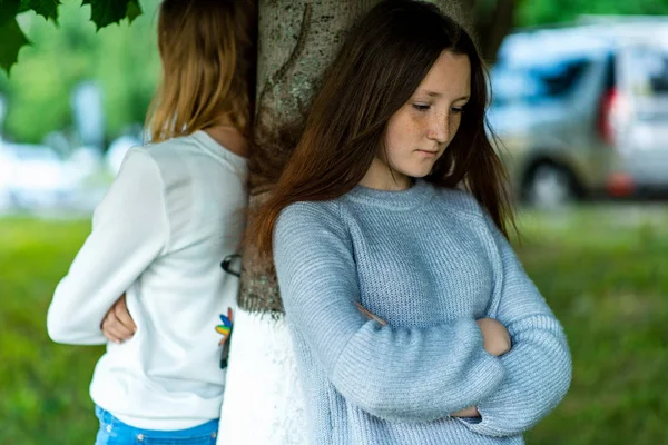 Two girlfriends in the summer in a park in nature. Offended each other. Friends quarreled. Negative among the girls. Problems in the relationship. Dissatisfaction with each other. Stand near a tree. — Stock Photo, Image