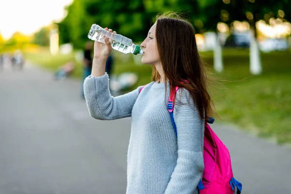 Красивая школьница. Летняя природа. Он держит бутылку воды и напитки. Брюнетка с длинными волосами. За спиной школьный рюкзак с учебниками. Зеленая улица в городе . — стоковое фото