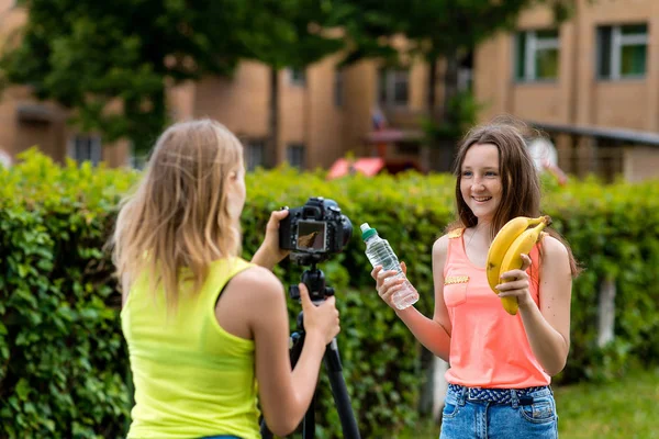 Dos chicas jóvenes. Verano en la naturaleza. Habla de una nutrición adecuada. En manos de una botella de agua y plátanos. Registrar suscriptores vlog y blog. Grabar lecciones de vídeo para Internet. Utilice la cámara con trípode . — Foto de Stock