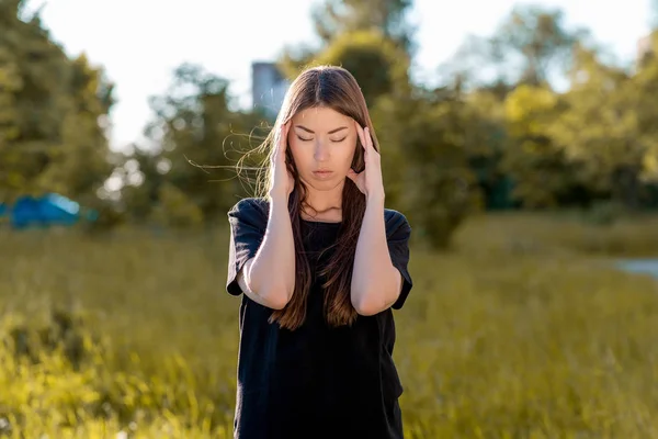 Ragazza bruna all'aria aperta. Tiene le mani intorno alla testa. Mal di testa. Cerca di concentrarsi. Emicrania e forte dolore. In estate nel parco vestito di coagulazione casuale . — Foto Stock