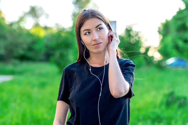 美しいブルネットの少女は、屋外の公園で夏にヘッドフォンで音楽を聴きます。感情的な空想する夢を見るします。良い笑顔の肯定的な思考 — ストック写真