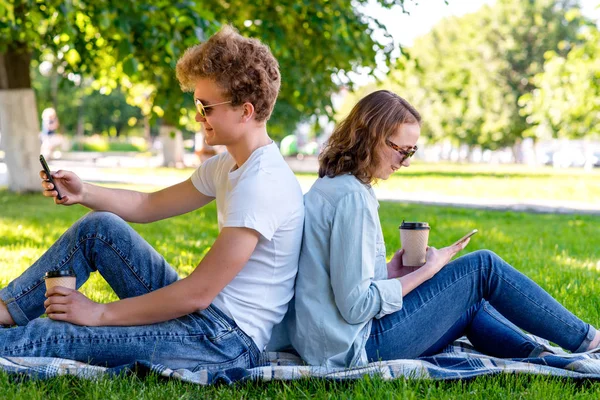 Studenci są facet z dziewczyna. Latem w parku. Posiada on herbaty w jego ręce. On ma odpoczynku po szkole Instytutu. Patrzy na smartfonie. Odpowiadać w sieciach społecznościowych. — Zdjęcie stockowe