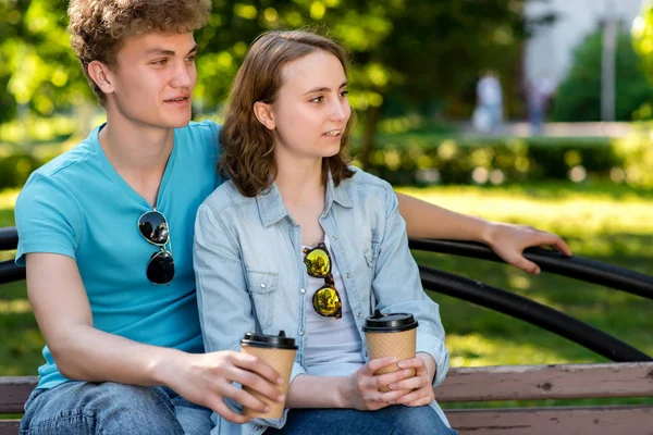 Mladý pár, studenty chlapec a dívka. Sedí na lavičce. V rukou drží šálky s kávou nebo čajem. Mluví a podívej se doprava. Ve skutečnosti před námi. Detail. — Stock fotografie