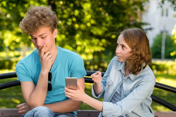 Une jeune fille pointe du doigt le gars du smartphone. Le problème est dans la relation. Des émotions de méfiance. Concepts de trahison et de trahison. Jeune couple en été dans un parc en plein air . — Photo