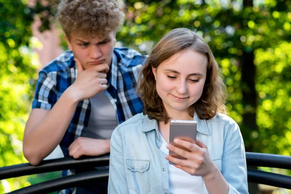 Krásná brunetka v letním parku na přírodu. V rukou drží smartphone komunikuje v sociálních sítích. Mladý muž špionů na její sms. Koncept nedůvěra k problému ve vztahu. — Stock fotografie