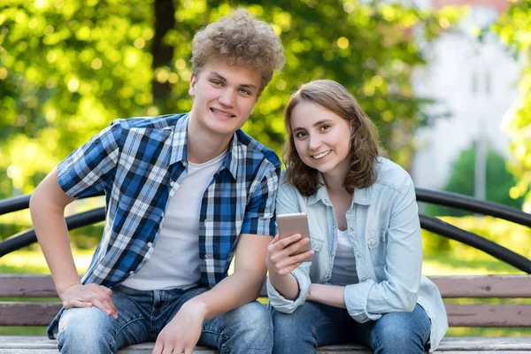 Ragazzo giovane coppia ragazza. Estate al parco su una panchina. Nelle sue mani tiene uno smartphone. Buon primo piano sorridente. Emozioni di felicità e piacere. Lo studente riposa dopo la scuola . — Foto Stock