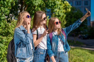 Park kız kız öğrenciler yaz aylarında. O kendini telefon çekiyor. Mutlu gülümseyerek poz fotoğraf makinesinde. Denim giysiler içinde. Kız öğrencilerin güneş gözlüğü takıyor. Açık havada şehirde.
