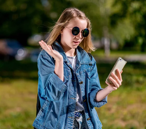 Colegiala en verano en el parque después de la escuela. En sus manos tiene un smartphone. Gestos de comunicación en una videollamada móvil. Vestido con ropa vaquera con gafas de sol. Al aire libre . —  Fotos de Stock