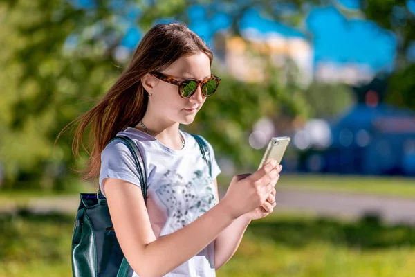 Gadis berambut cokelat yang cantik. Di musim panas di taman. Di tangannya memegang smartphone. Menulis pesan di jejaring sosial. Dengan kaos putih dan kacamata hitam. Istirahat setelah sekolah . — Stok Foto
