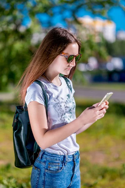 Mooie brunette meisje. In de zomer in het park. In zijn handen houdt een smartphone. Schrijft berichten op sociale netwerken. In een wit T-shirt en zonnebril. Rusten na school. — Stockfoto