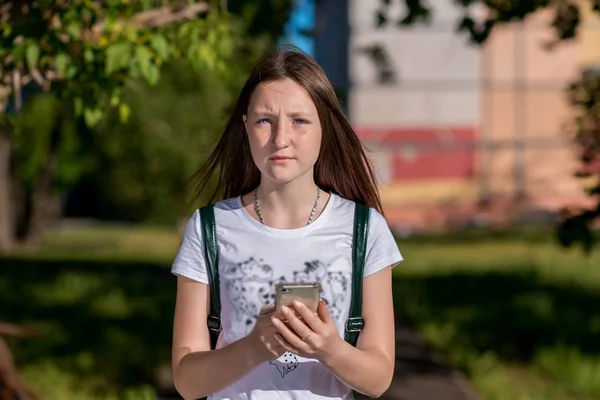 Girl Brunette Schoolgirl. In summer in the city in nature. In his hands holds a smartphone. The concept of a call to parents. Emotionally looks into the frame. He rest after school. — Stock Photo, Image