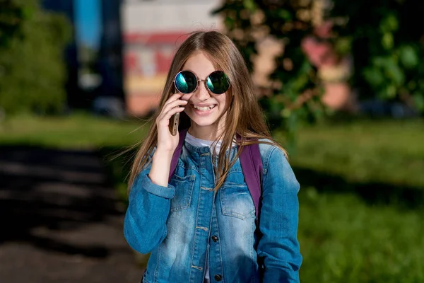 Gadis sekolahan. Musim panas di alam. Di tangan memegang smartphone berkomunikasi melalui telepon. Senyum dengan gembira. Dalam kacamata hitam dan jaket denim. Emosi sukacita dan kesenangan. Konsep kehidupan positif . — Stok Foto
