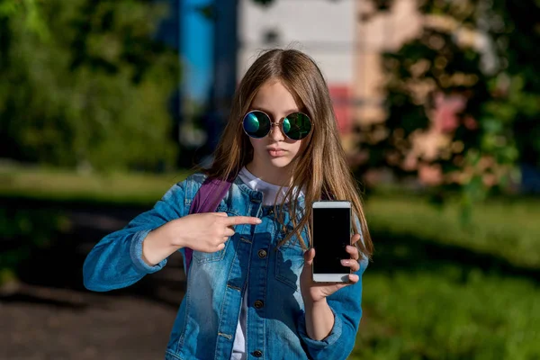 Colegiala en gafas de sol. Verano en la naturaleza. En sus manos tiene un smartphone. Un dedo señala el teléfono. El concepto es una nueva aplicación de gadget. Concentración del foco de emoción . —  Fotos de Stock