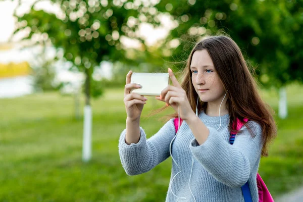 Όμορφη μαθήτρια κορίτσι. Το καλοκαίρι στο πάρκο στη φύση. Στα χέρια του κρατά ένα smartphone. Πάρει τις εικόνες του τοπίου. Ελεύθερος χώρος για το κείμενο. Να χαμογελά με φακίδες στο πρόσωπό της. Ανάπαυση μετά το σχολείο. — Φωτογραφία Αρχείου