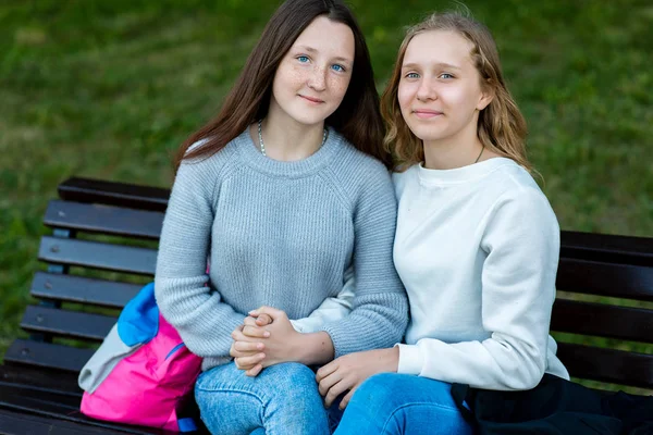 Dos colegialas en verano en un banco. Camping después de la escuela. Se toman de las manos. El concepto de amistad son las mejores amigas. Una sonrisa feliz es disfrutar. Mirada emocional . — Foto de Stock