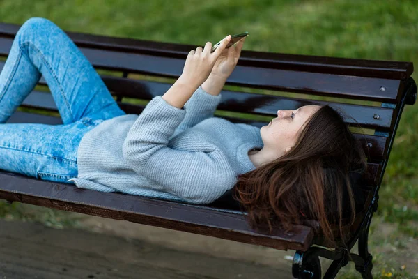 Девочка-школьница лежит на скамейке. В его руках смартфон. Просмотр видео читает сообщения в социальных сетях. Концепция детей в Интернете. Эмоции наслаждаясь отдыхом на природе . — стоковое фото