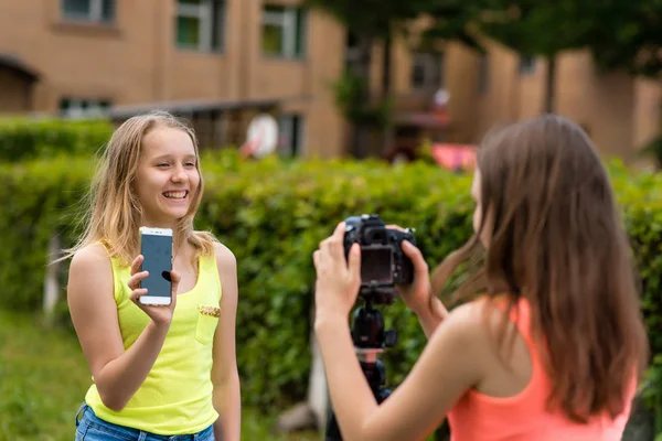 Dva přátelé dívka. Letní přírody. Píše video kameru. Ve svých rukou drží smartphone. Koncept mladých bloggerů je nahrávání videa pro Internet. Emoční se šťastně usmívá. Hovořit o miniaplikace. — Stock fotografie