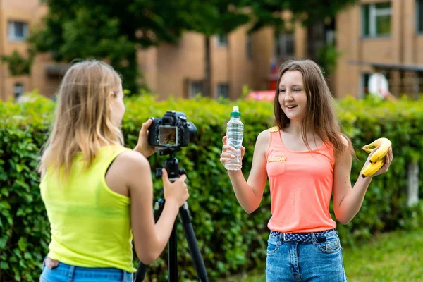 Δύο φίλες. Το καλοκαίρι στη φύση. Εγγραφή βίντεο στη φωτογραφική μηχανή. Η έννοια του νέους bloggers. Υγιεινή διατροφή. Συναισθήματα μιλούν στην κάμερα. Στα χέρια της κρατά ένα μπουκάλι νερό και μπανάνες. — Φωτογραφία Αρχείου
