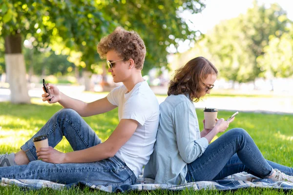 Una giovane coppia. Estate nella natura. Un ragazzo con una ragazza che riposa su un plaid. Contiene caffè caldo o tè. Guardano gli smartphone. Scrivi messaggi sui social network . — Foto Stock