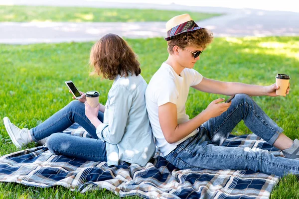 Kluk s holkou v létě relaxovat v přírodě. Je držitelem smartphone ve svých rukou a čte korespondenci. Ve svých rukou drží sklenku kávy nebo čaje. Se těší relaxační přehoz. — Stock fotografie