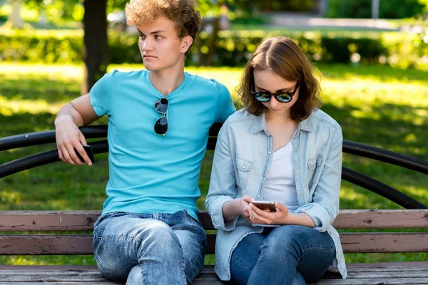 Un tipo con una chica en verano en un parque en la naturaleza. Se sientan en un banco. En manos de un smartphone está leyendo una red social. Resto de estudiantes en la naturaleza . —  Fotos de Stock