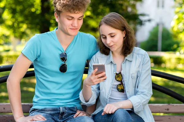 Un chico y una chica están sentados en un banco. Verano en la naturaleza. Chica en las manos sosteniendo un smartphone. Emocionalmente sonriente lee una red social . —  Fotos de Stock