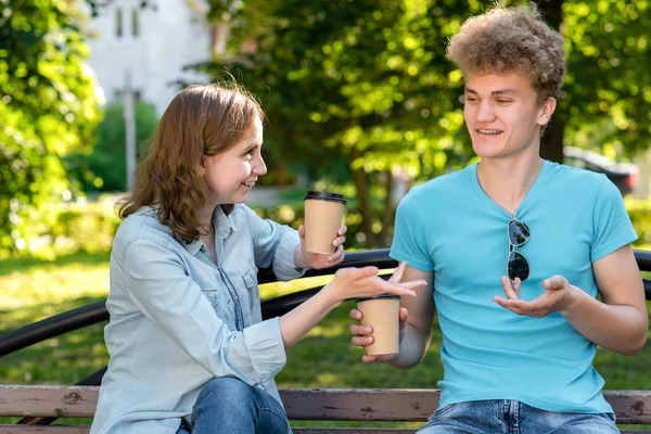 Ein Kerl mit einem glücklichen Mädchen in einem Park in der Natur. Im Sommer sitzen sie auf der Bank. Er hält Tassen mit Kaffee oder Tee in der Hand. er gestikuliert emotional mit seinen Händen. — Stockfoto