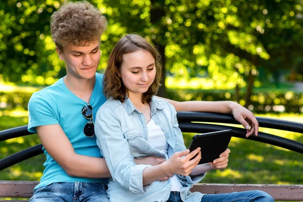 Mladí studenti v létě v parku venku. Sedí na lavičce v městě. Ten chlap je objímat dívku. Dívka v rukou drží tablet. Usmíval se spokojeně sledovat video na internetu. — Stock fotografie