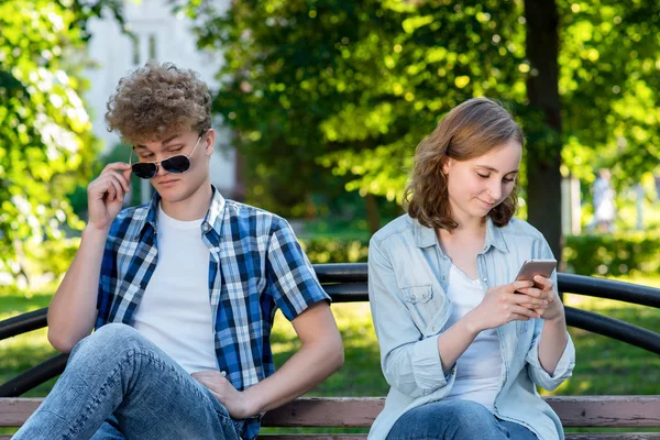 The guy and the girl are sitting on bench. Summer in nature. A girl in her hands holds a smartphone communicates in social networks. The guy spies Through glasses behind the girl.